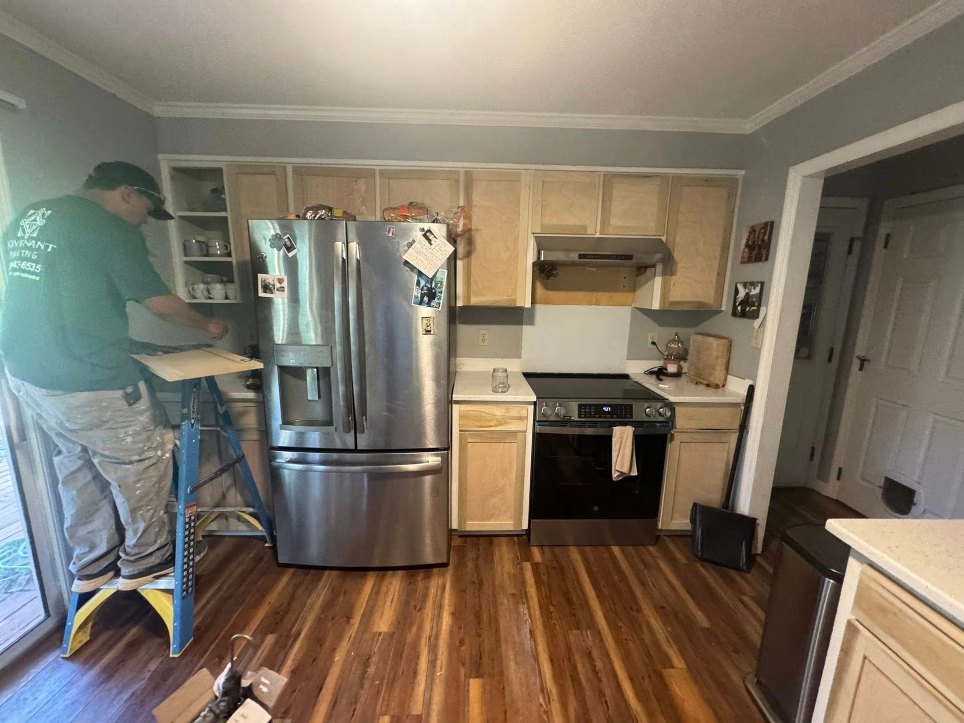 Man on ladder working on kitchen cabinets; stainless steel appliances, wooden floor.