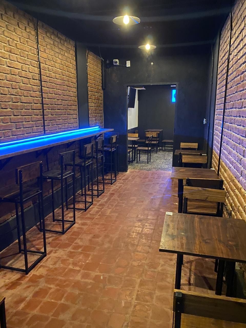 Un estrecho bar con paredes de ladrillo, barra iluminada con neón azul, taburetes altos y mesas de madera bajo luces tenues.