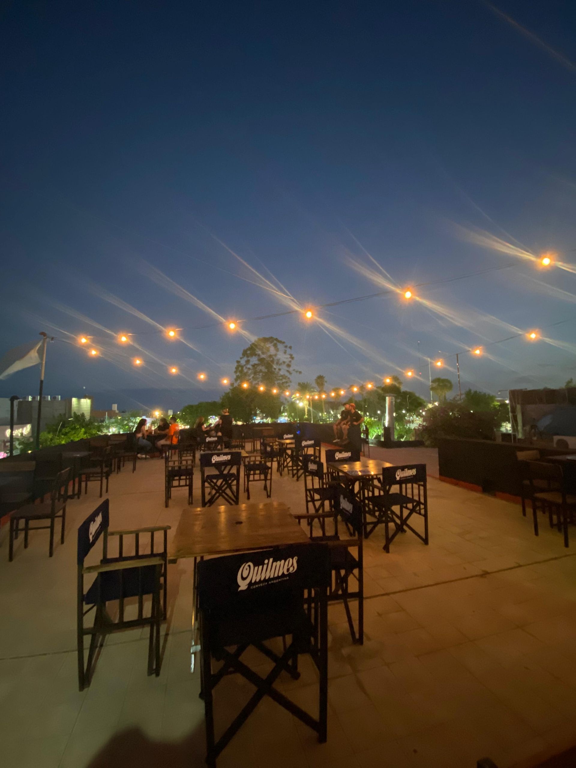 Comedor al aire libre en un patio con luces de guirnalda y sillas negras sobre una terraza de hormigón, al atardecer.