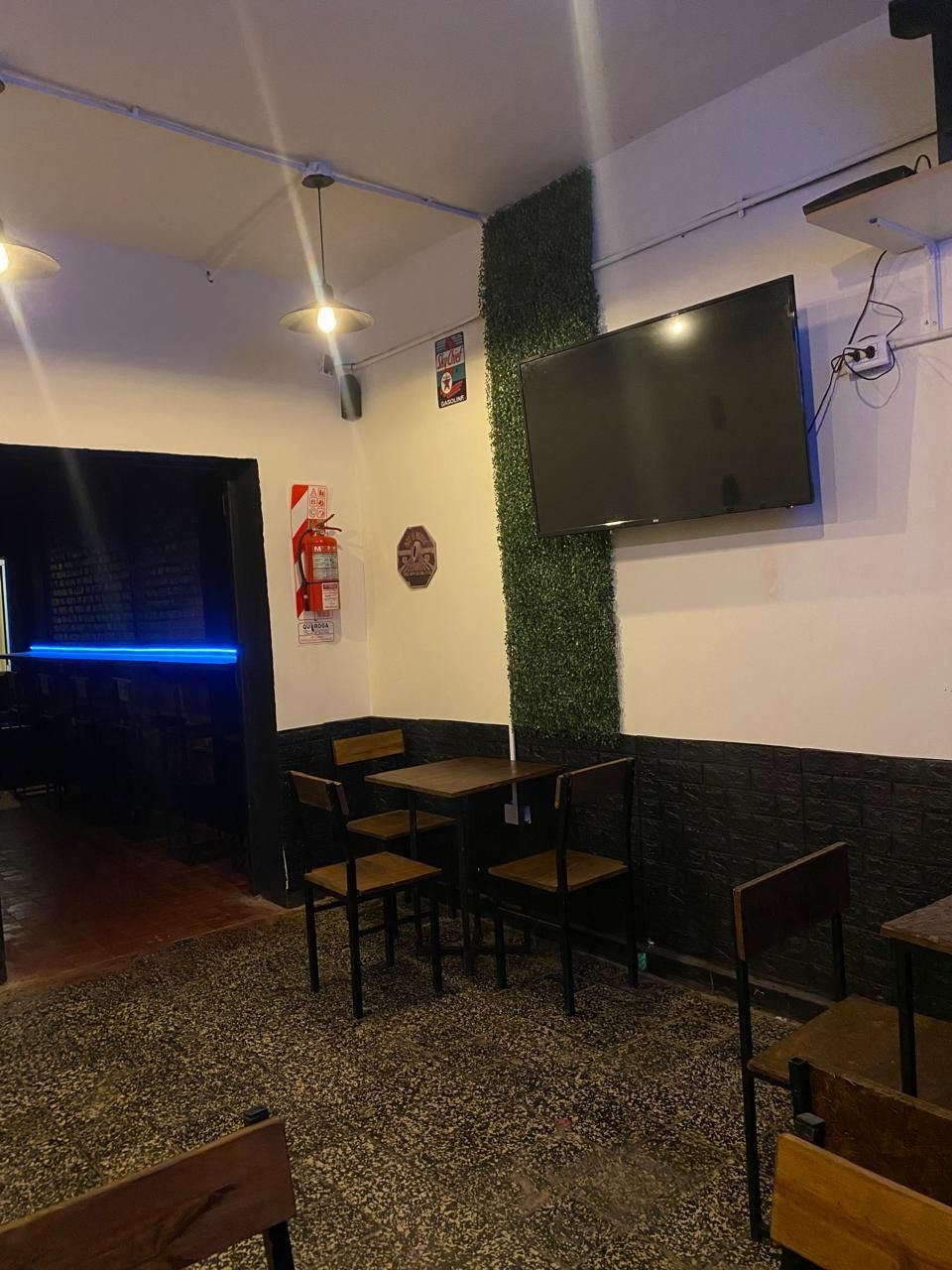 Interior de una pequeña cafetería con mesas, sillas, televisor de pared y una puerta iluminada con luz azul.