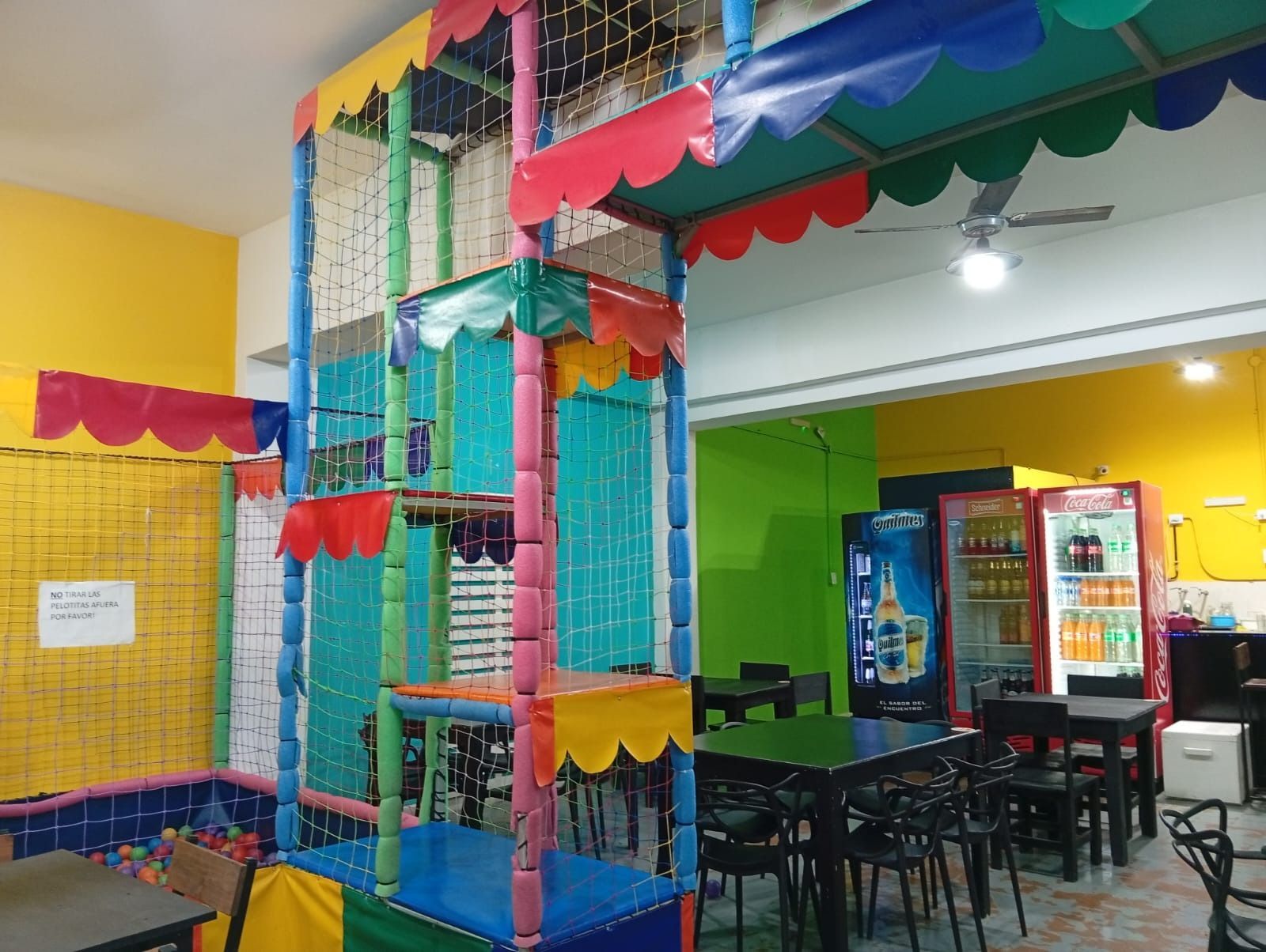 Zona de juegos interior colorida con una estructura alta para trepar, túneles de malla y mesas en una habitación luminosa.