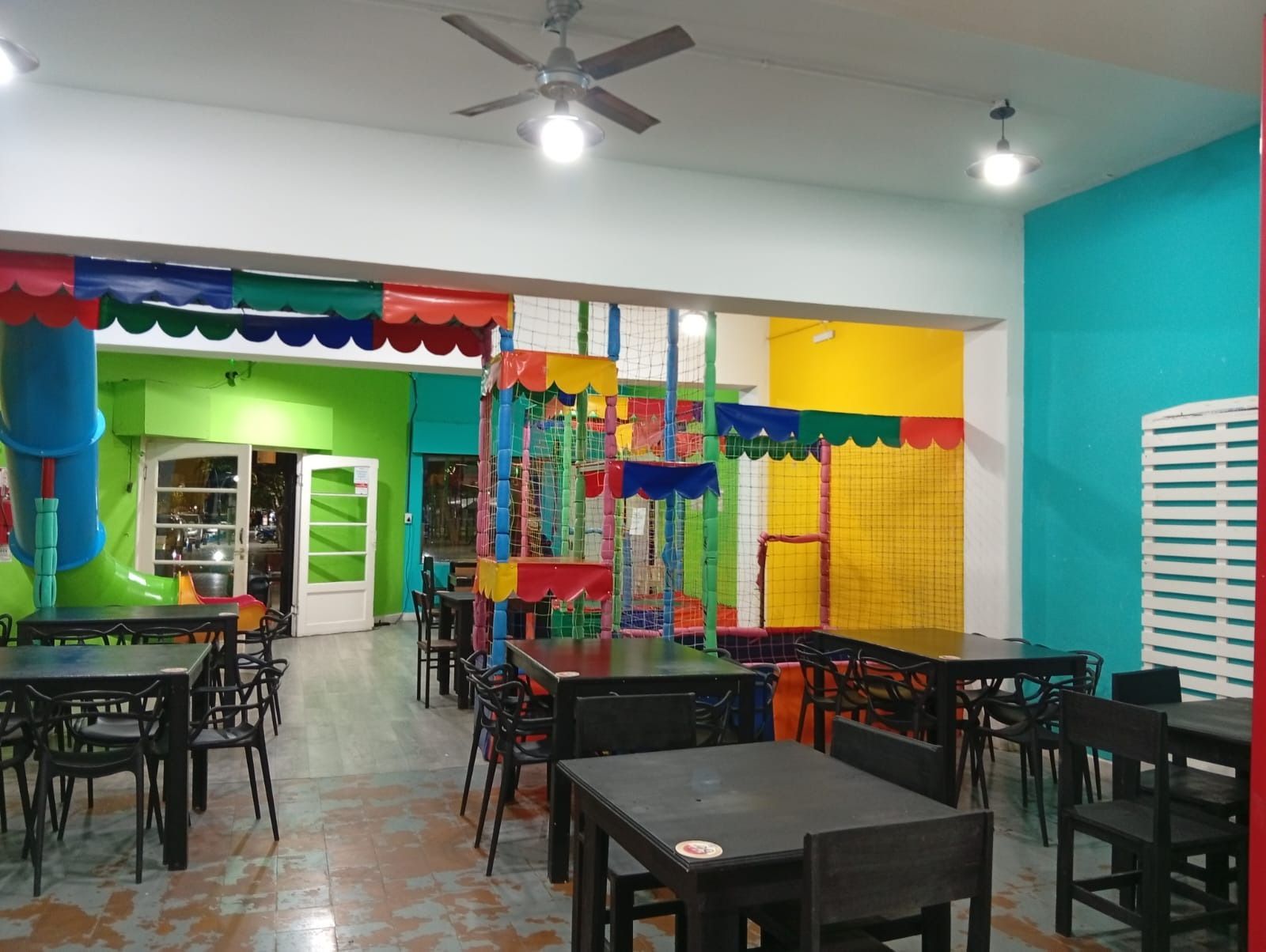 Comedor interior colorido con mesas y sillas negras, paredes turquesas y zona de juegos decorativa en la parte trasera.