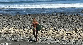 A woman in a bikini is walking on a rocky beach with a surfboard.