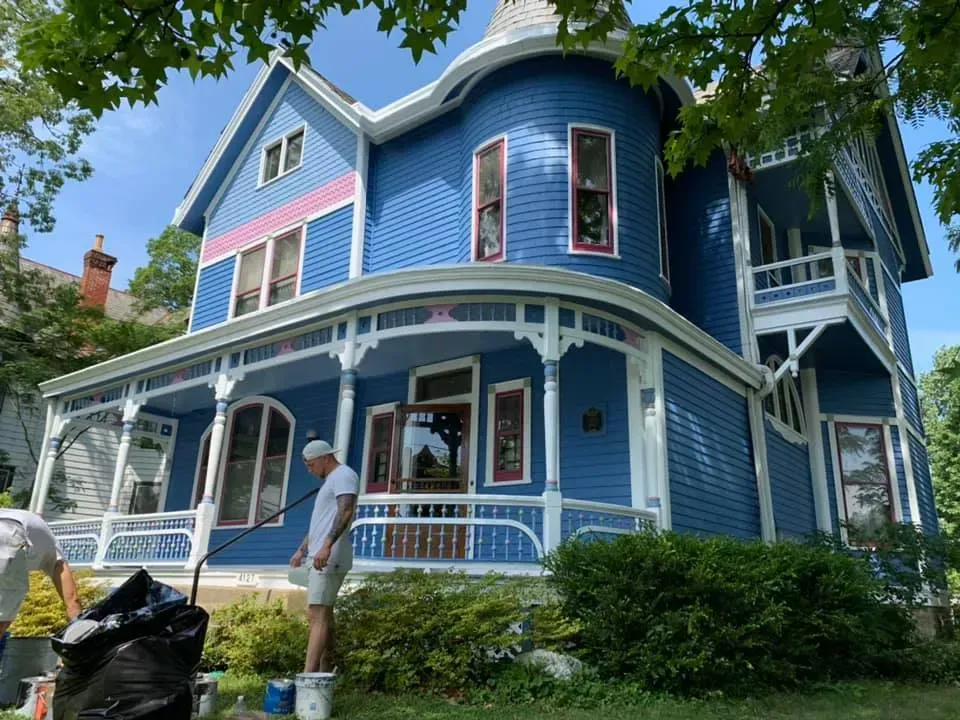 A man is painting a blue house with white trim.