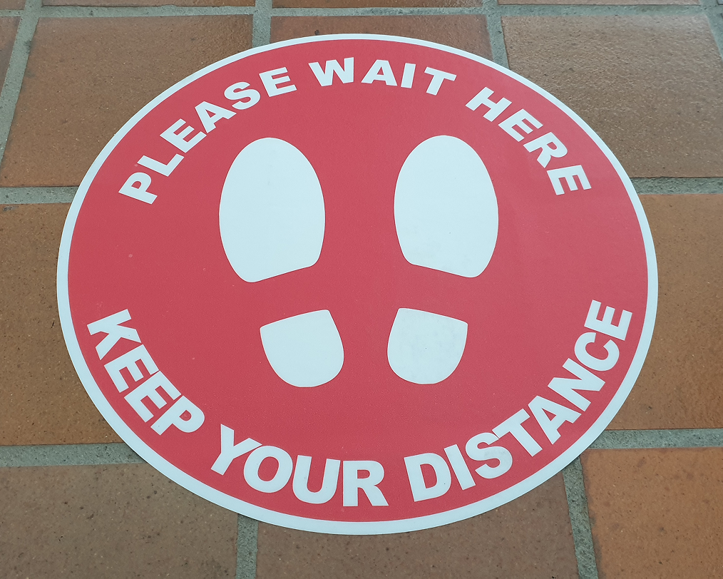 A red and white sign that says please wait here keep your distance A red and white sign that says please wait here keep your distance