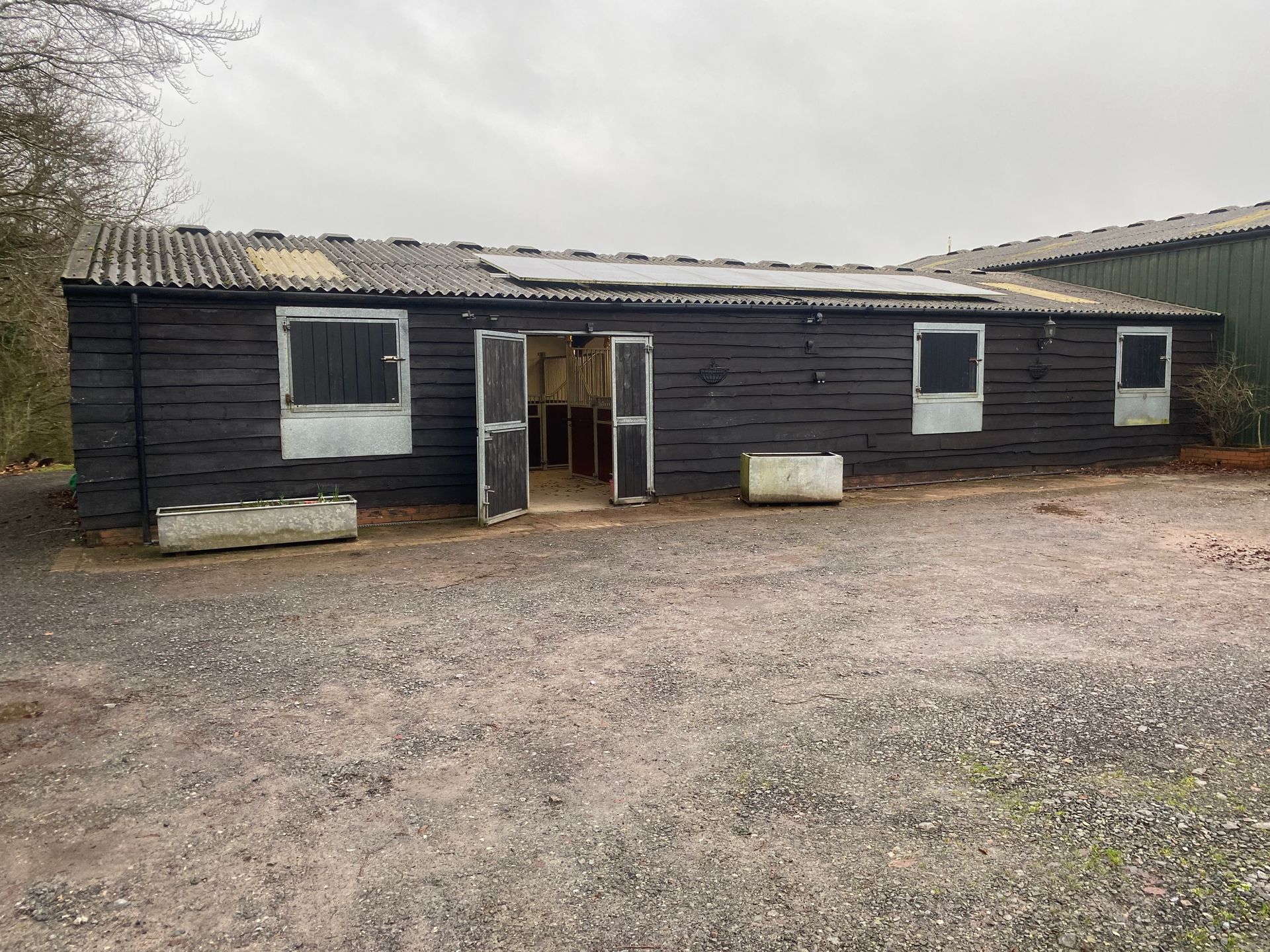Black shed with open doors, windows, and weathered roof on a gravel surface under an overcast sky.