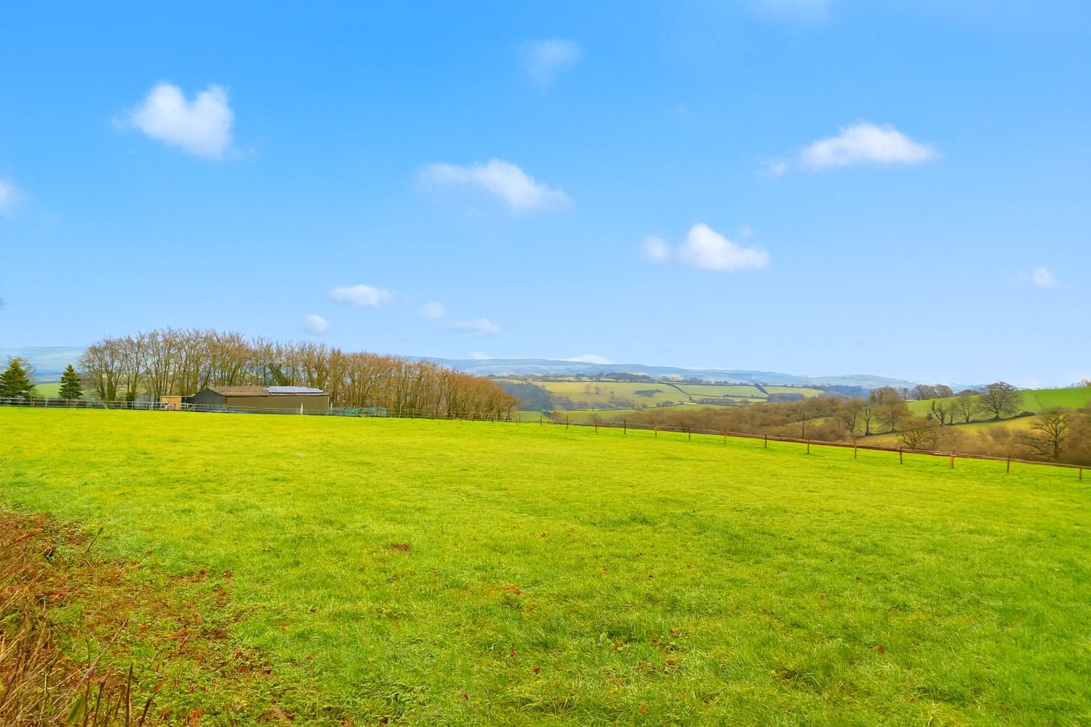 Green field under blue sky with sparse clouds, a small building and distant hills.