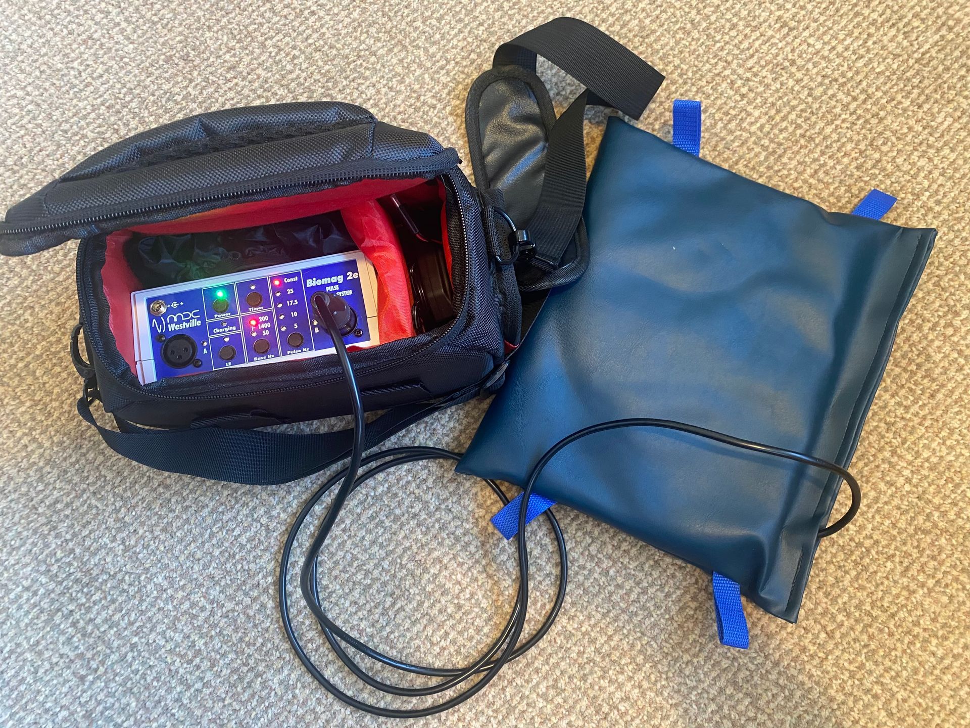 Black camera bag with a device and a blue pouch, connected by a black cord, on a neutral-colored surface.