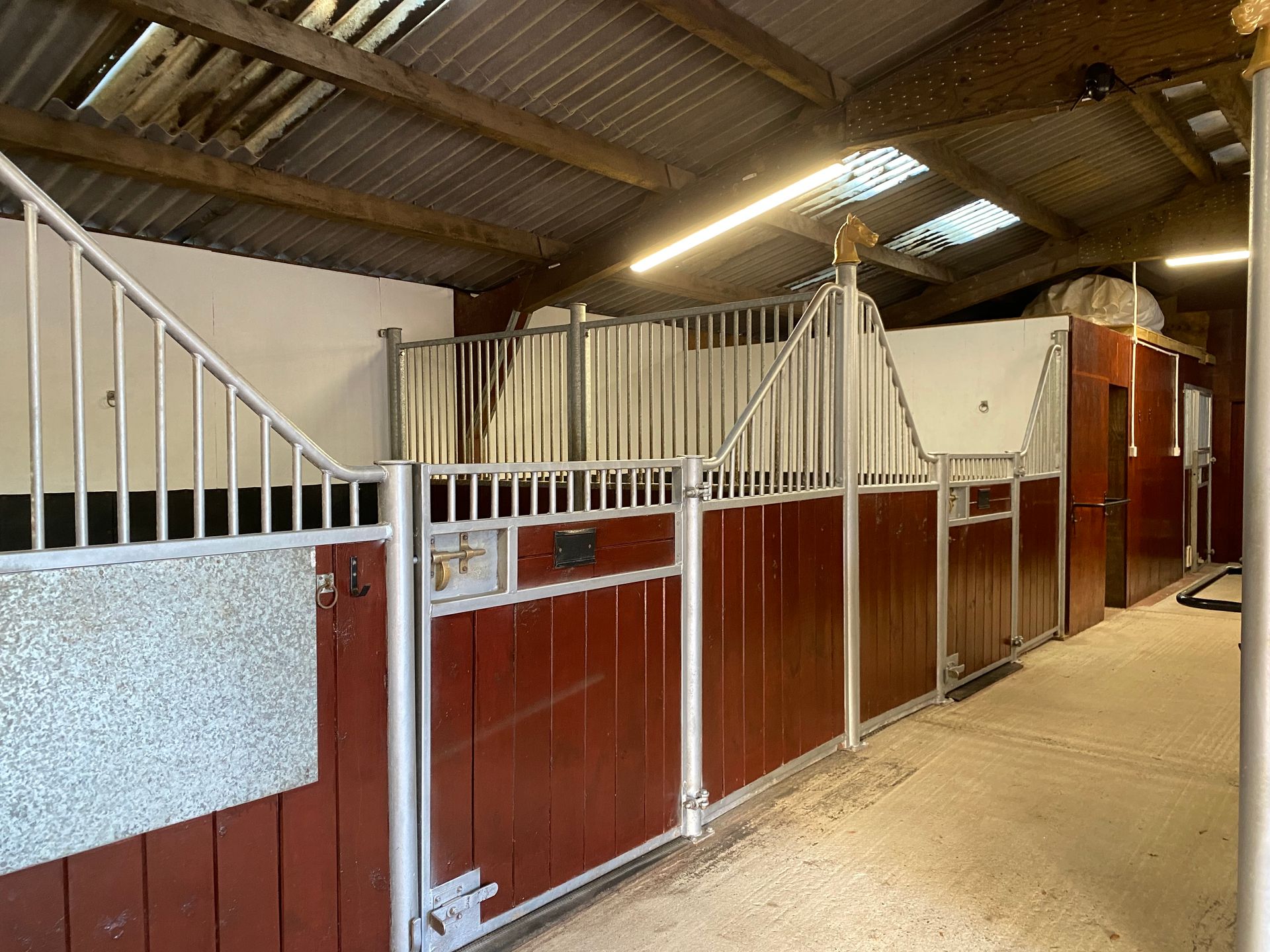 Horse stable with red-painted wooden stall fronts, metal bars, and a concrete floor.