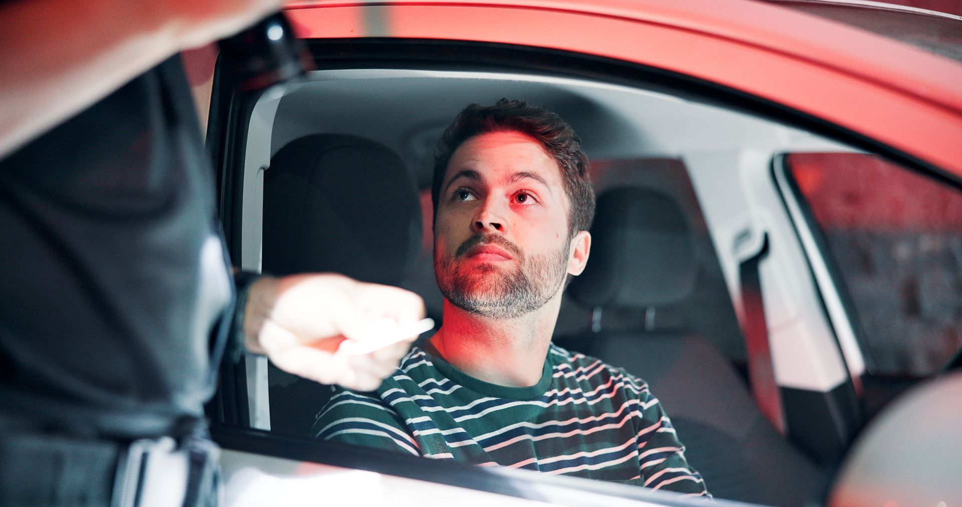 Man in car being handed a test strip by a police officer, lit by red emergency lights.