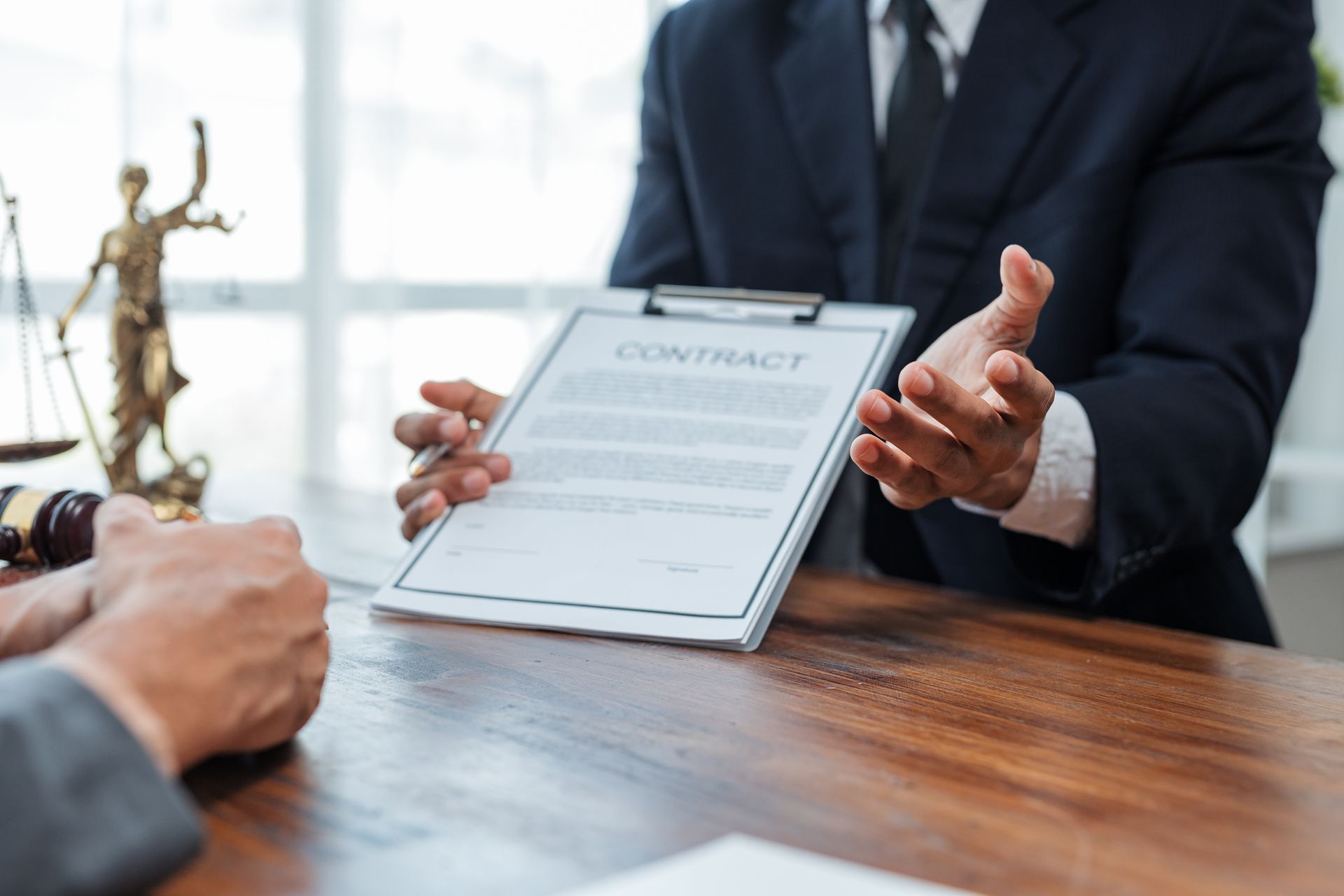 Person in suit presenting a contract to another person at a wooden table in an office, a scale of justice statue visible.