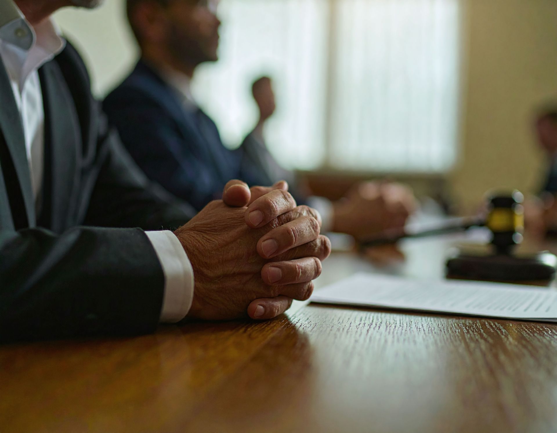 Close-up of hands clasped on a table, with people in suits, papers, and a gavel in a meeting.