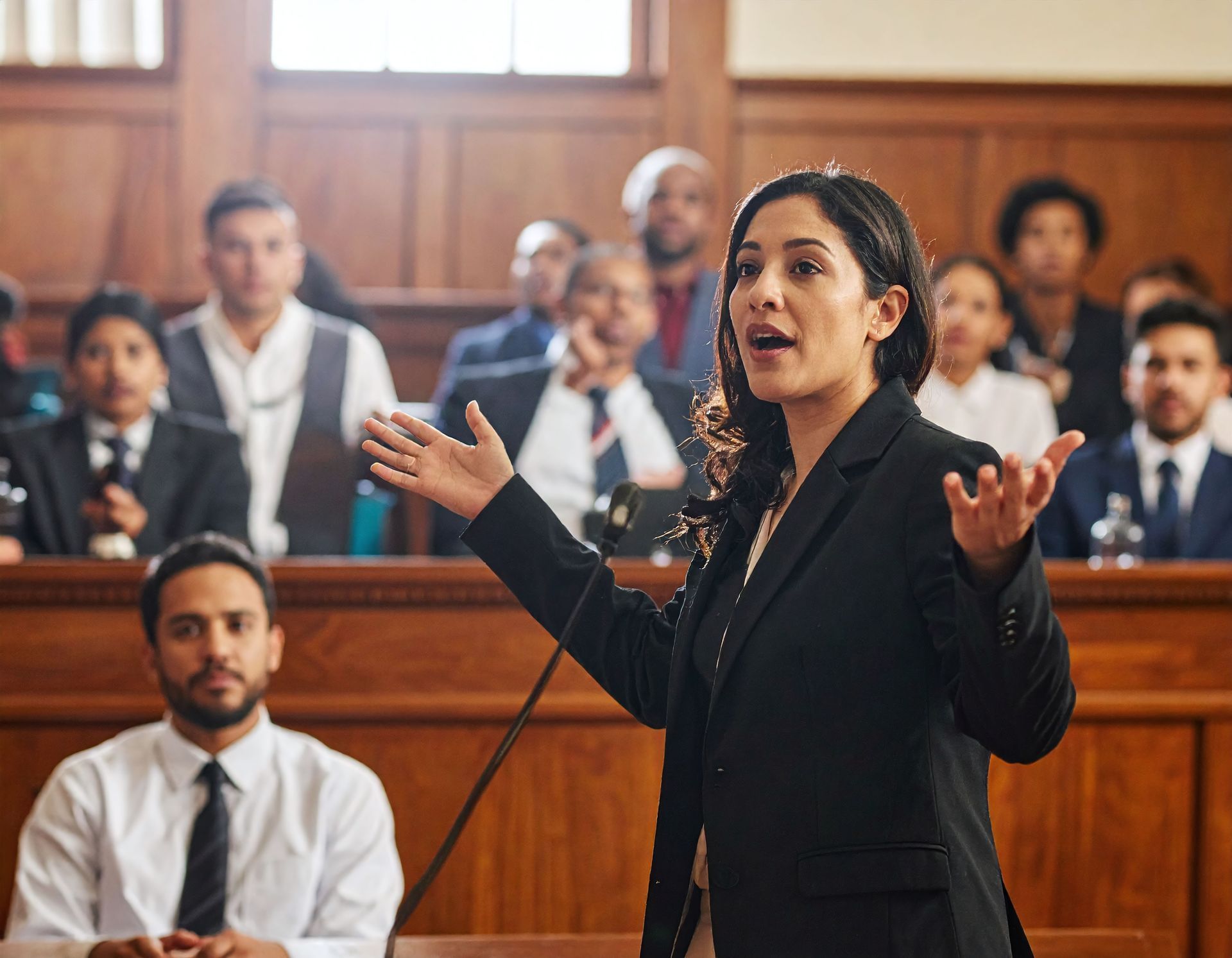 Woman in a courtroom speaking with hands raised; other people sit in the background.