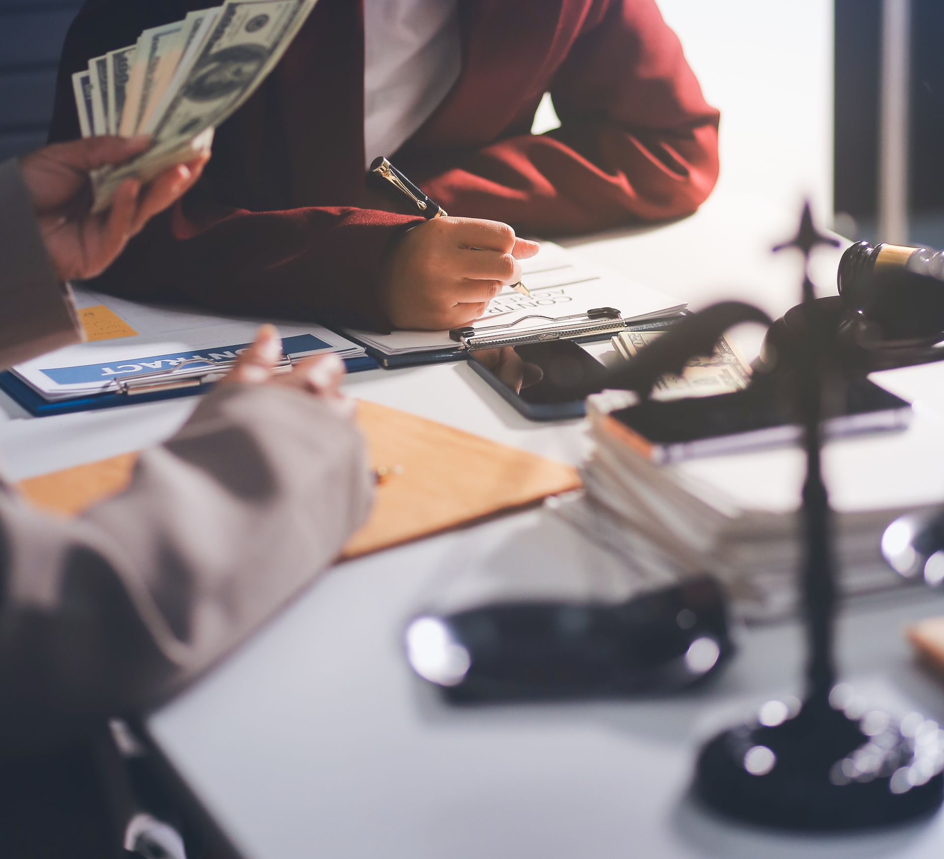 Person handing money to another person signing documents at a table; scales of justice in the background.