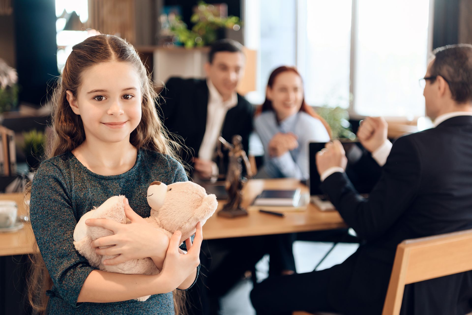 Girl holding teddy bear smiles in foreground; parents and lawyer in background, at a table.