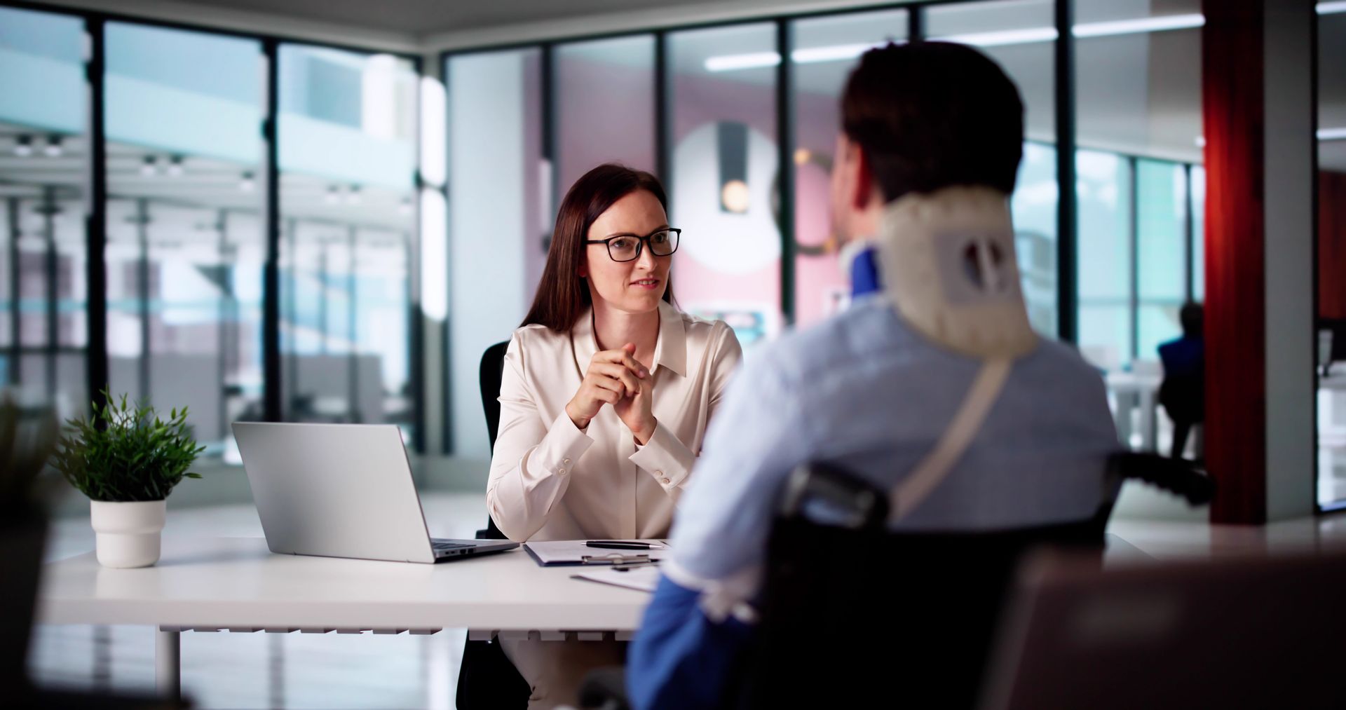 Woman in glasses at desk talking to person in wheelchair and neck brace. Office setting.