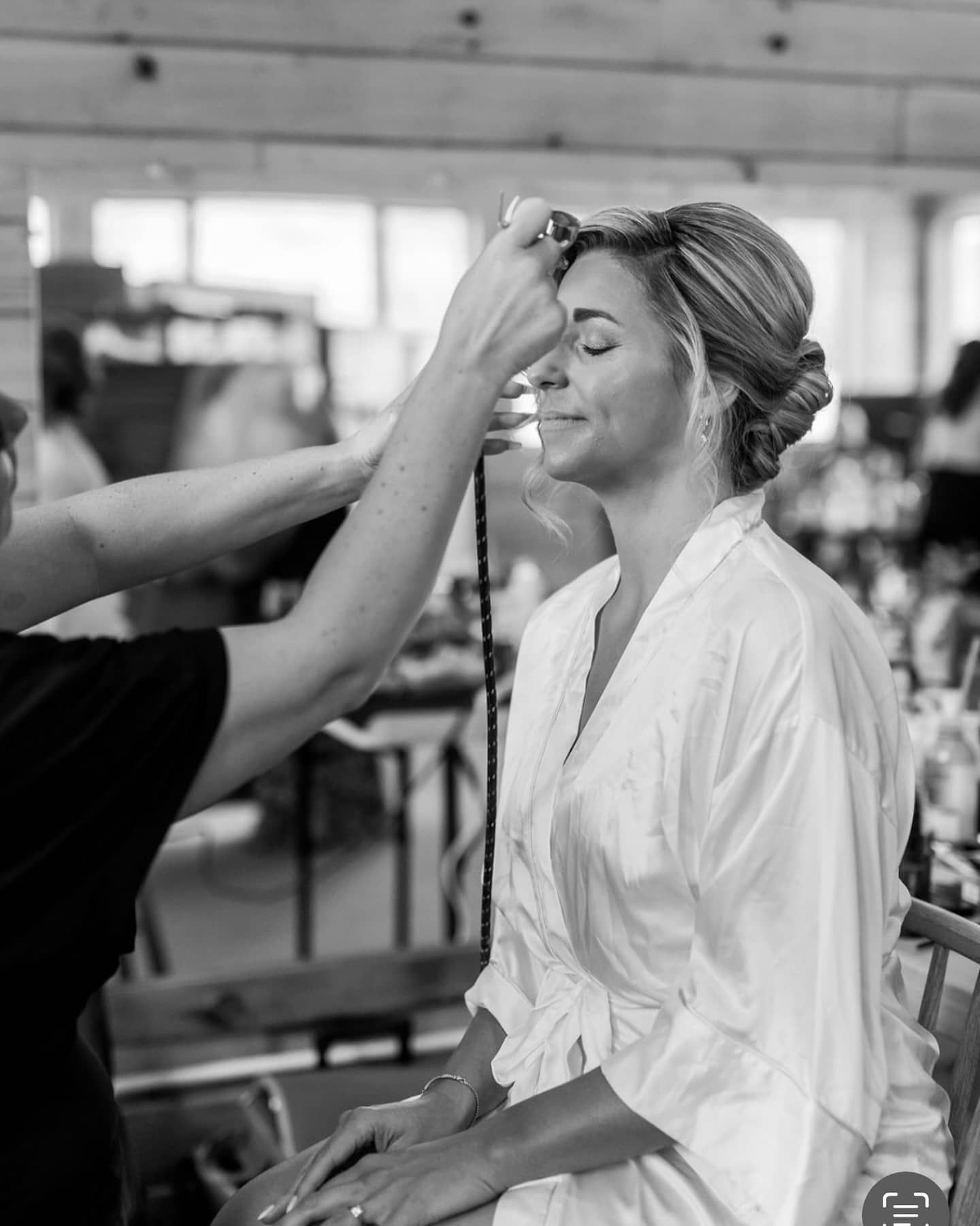 Bride having her face airbrushed before wedding, wearing a white robe, in a well-lit room.