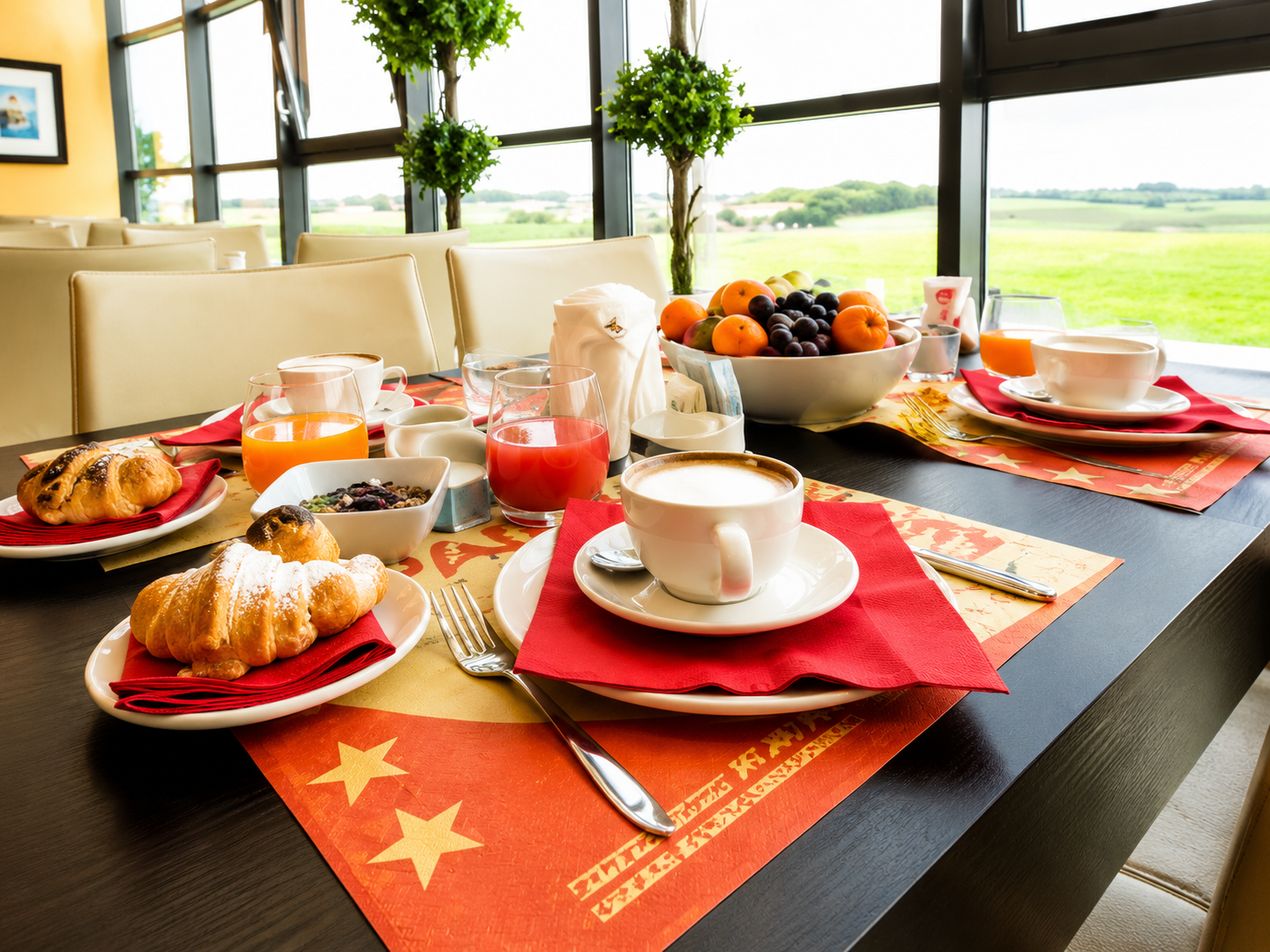 Tavolo della colazione apparecchiato con caffè, frutta, pasticcini e tovagliette arancioni accanto a una finestra luminosa