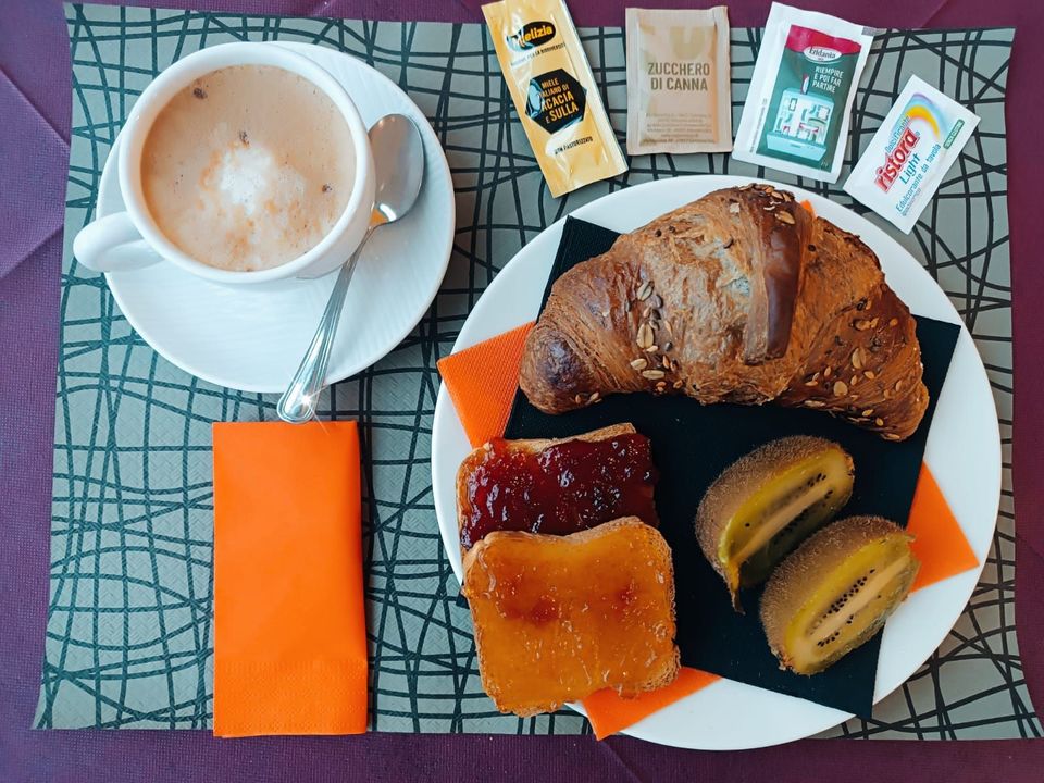 Tavolo della colazione apparecchiato con caffè, frutta, pasticcini e tovagliette arancioni accanto a una finestra luminosa