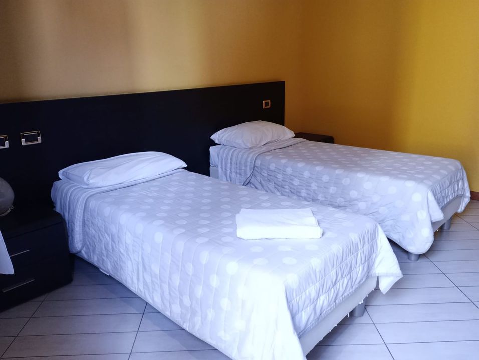 Due letti singoli con lenzuola bianche in una camera d'albergo beige, con testiera nera e pavimento piastrellato.