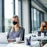 A woman wearing a mask is sitting at a desk in front of a laptop computer.