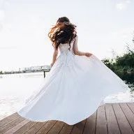 A woman in a white wedding dress is standing on a wooden dock.
