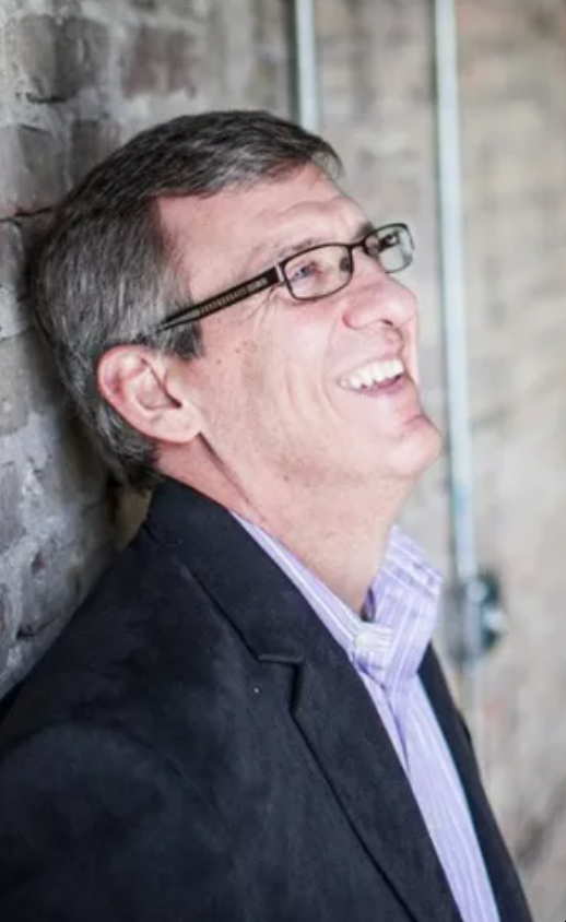 A man wearing glasses and a suit is leaning against a brick wall and smiling.