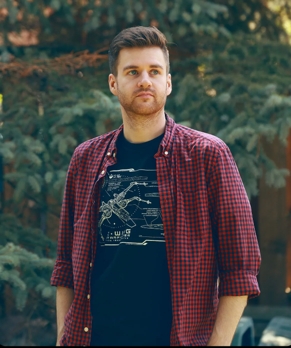 A man wearing a plaid shirt and a black t-shirt is standing in front of trees.