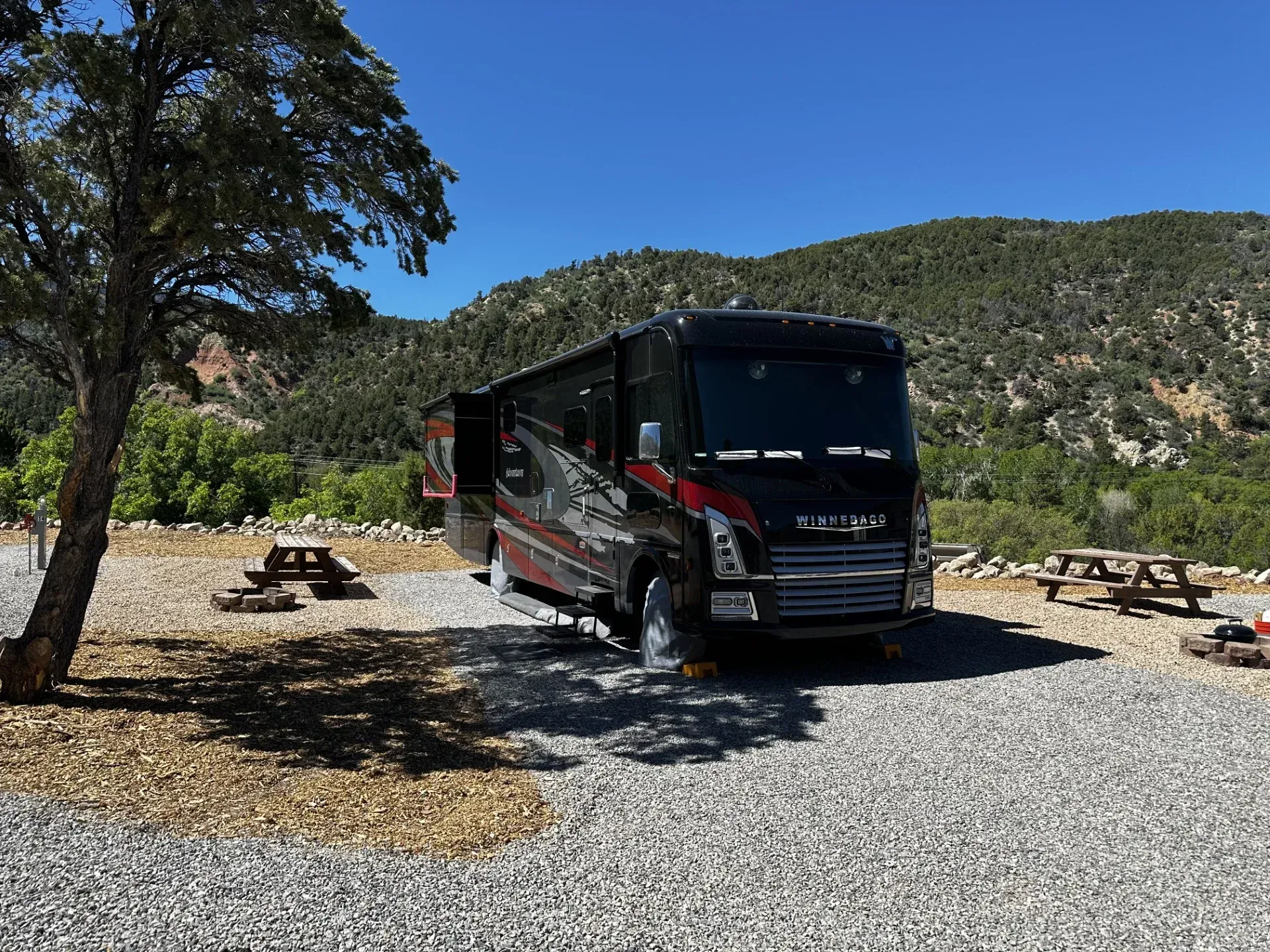 A large rv is parked in a gravel lot with mountains in the background.