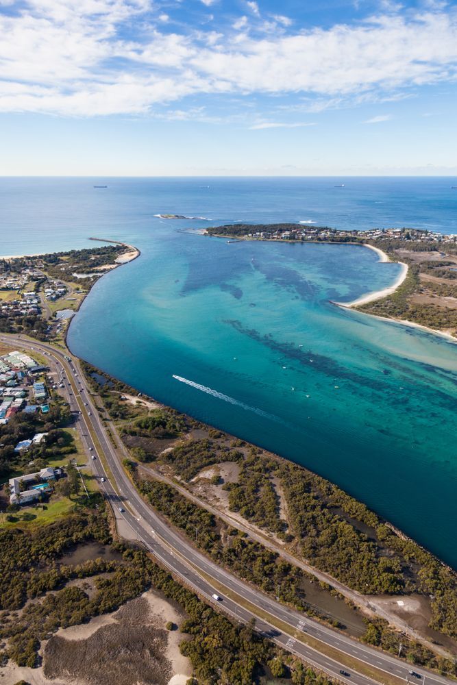 Aerial view of a bright blue channel meeting the ocean, with a road running alongside the channel through a coastal landscape. — GMS Legal & Conveyancing In Lake Macquarie, NSW