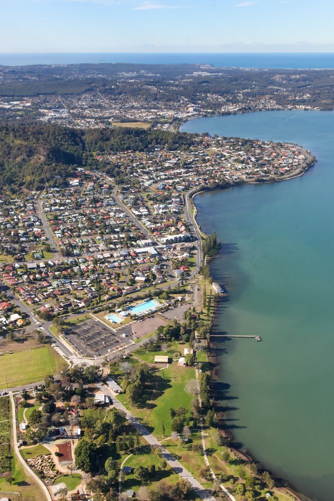Aerial view of a coastal town with buildings, a waterfront, and a pier extending into blue water. - GMS Legal & Conveyancing in Toukley NSW