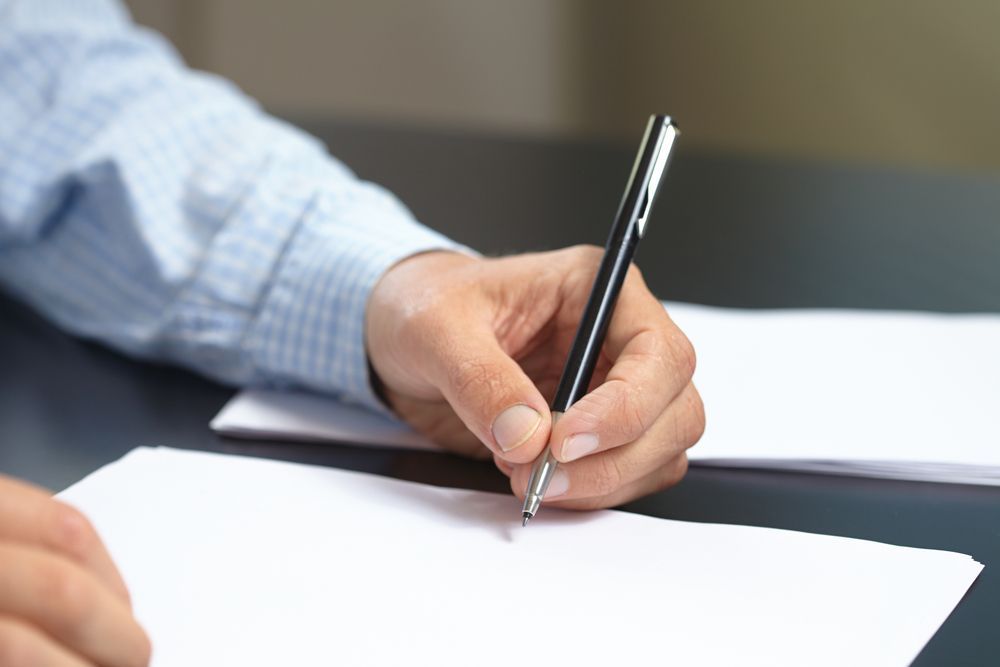Person's hand holding a pen writing on a blank white sheet of paper on a dark surface. GMS Legal & Conveyancing In 38 Canton Beach Rd, Toukley