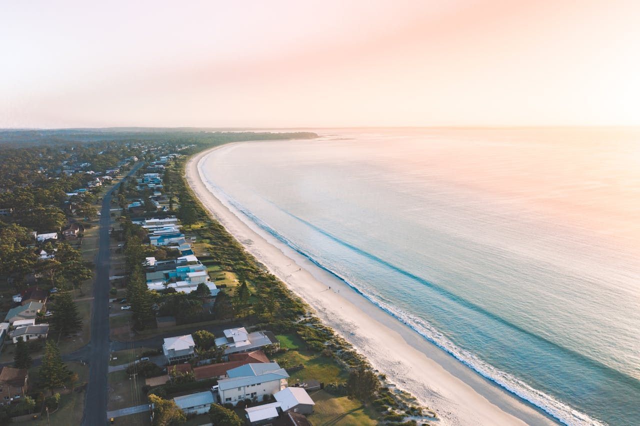 Aerial view of beach and coastline with calm, blue water and houses along the shore. GMS Legal & Conveyancing In 38 Canton Beach Rd Toukley, NSW