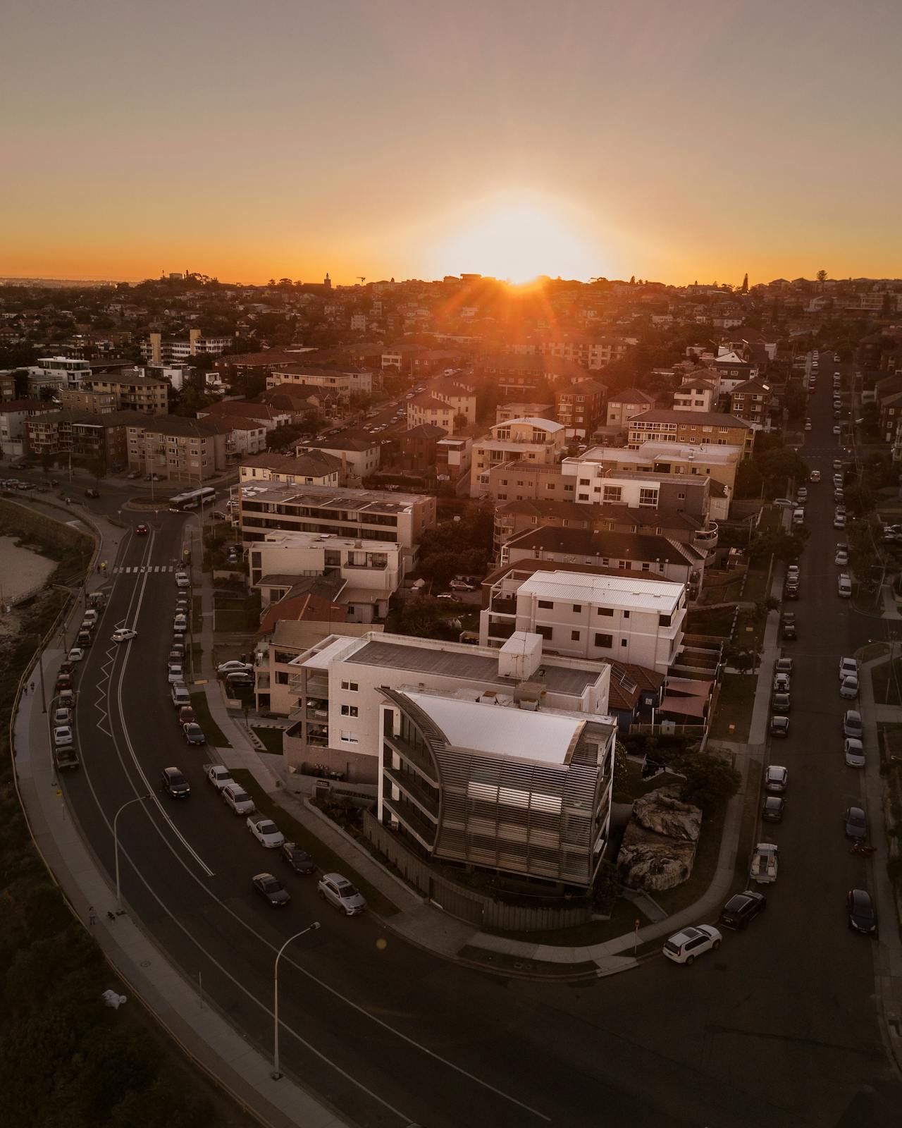 Sunset over a coastal city; modern white building, curving road lined with cars, houses. GMS Legal & Conveyancing In 38 Canton Beach Rd Toukley, NSW