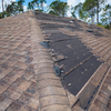 Damaged brown shingle roof with missing sections exposing the black underlayment; trees in the background.