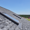 Gray shingle roof with three skylights against a blue sky and trees in the background.