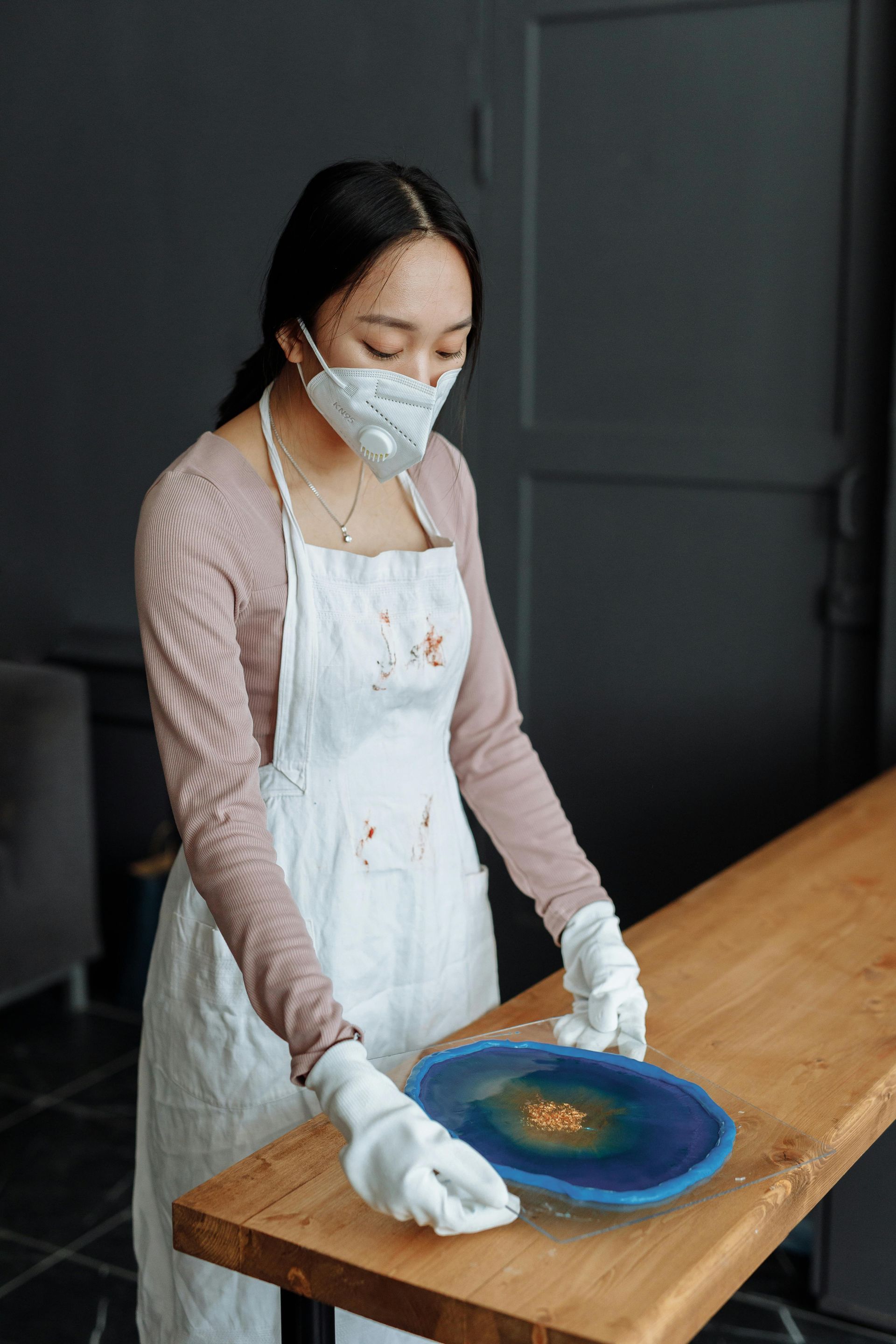 A woman wearing a mask and apron is cleaning a wooden table.
