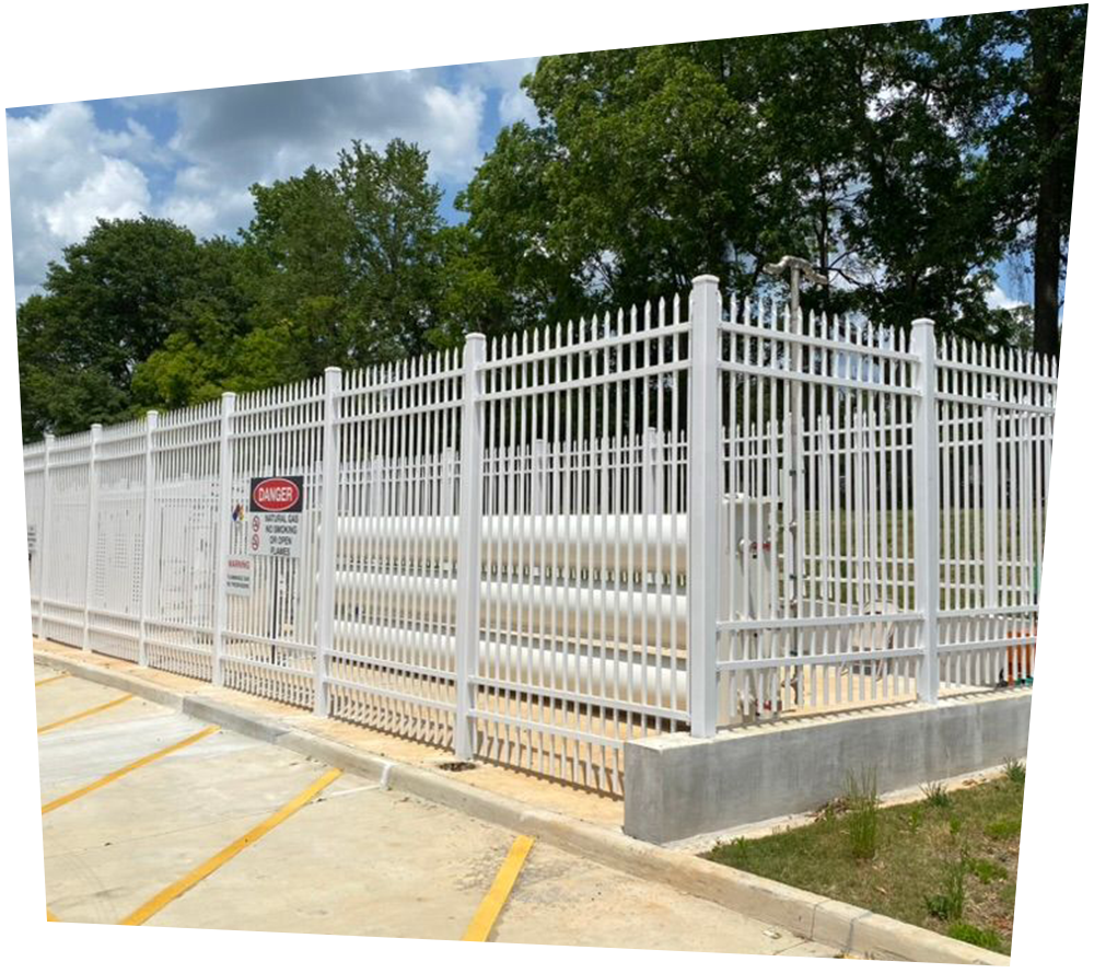 White fenced enclosure with pipelines, on a concrete base, near trees and a parking lot.