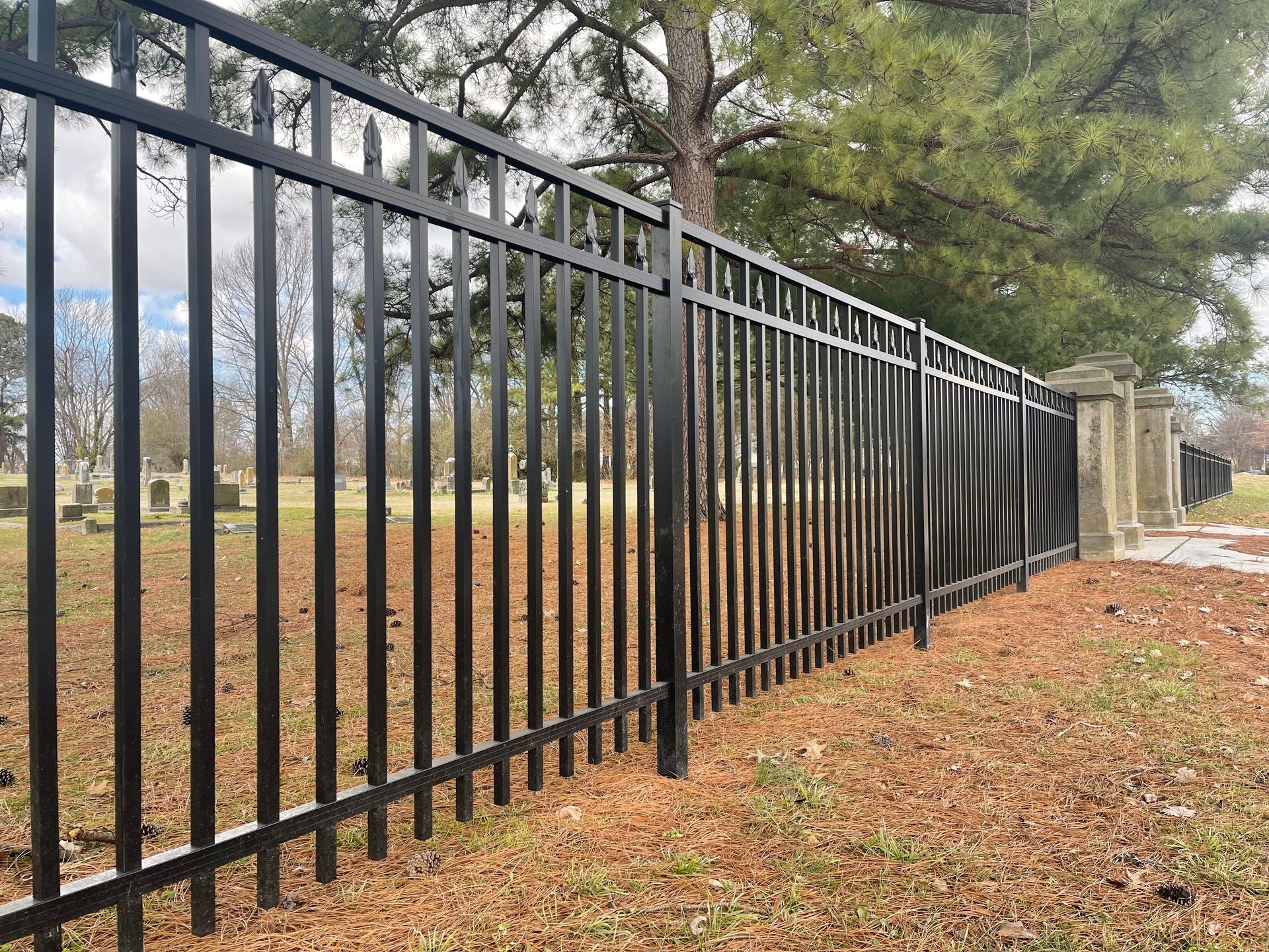 A black metal fence surrounds a cemetery with trees in the background.