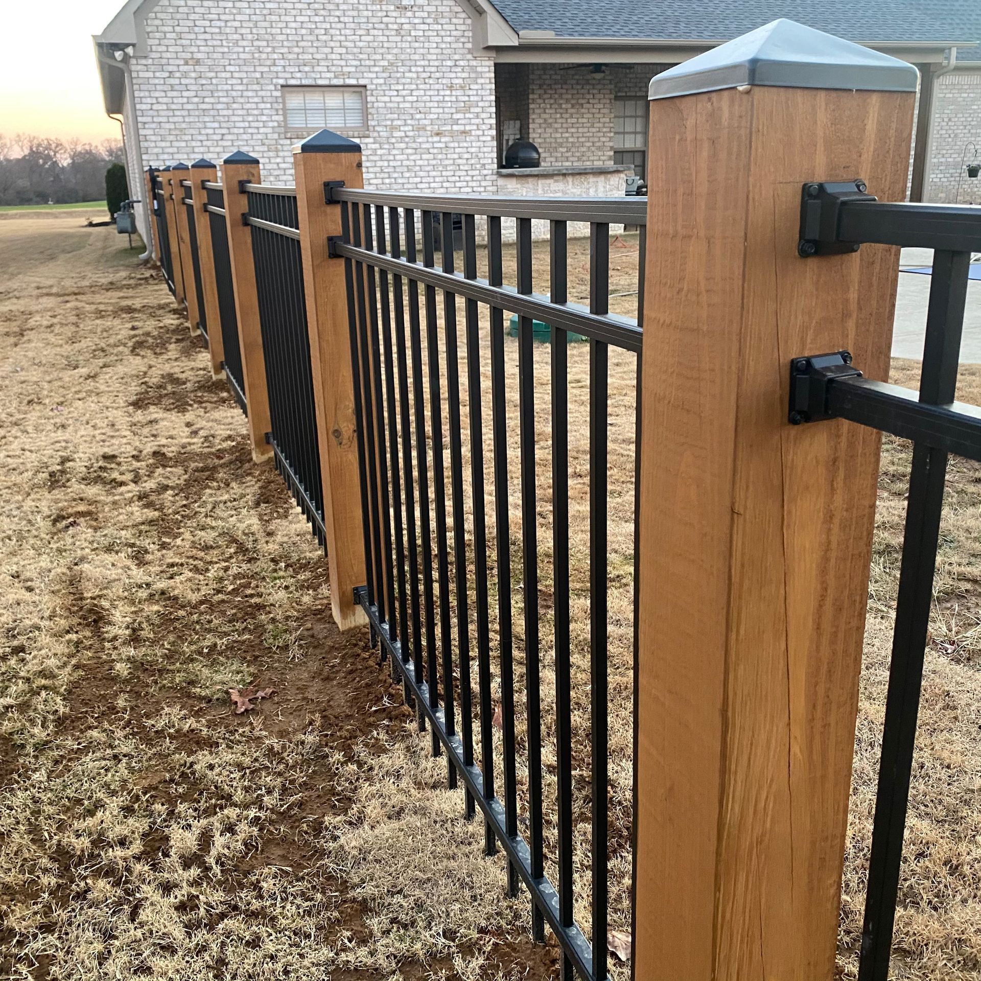 A metal fence with wooden posts is in front of a white brick house.