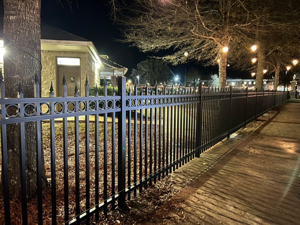 Black metal fence along a sidewalk at night, with trees and string lights.