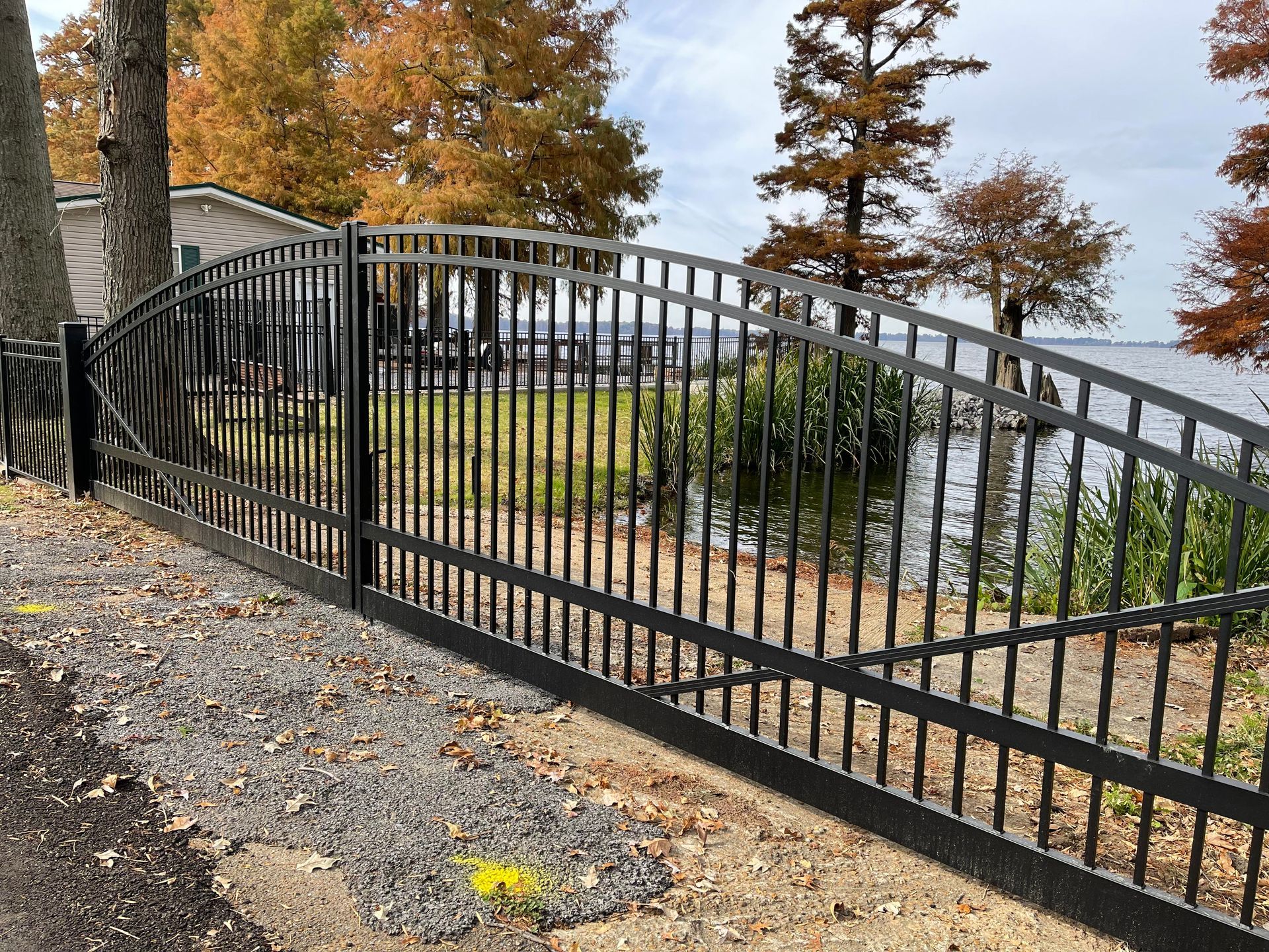 Black metal gate along a shoreline, with an arched top and vertical bars. Trees and water in the background.
