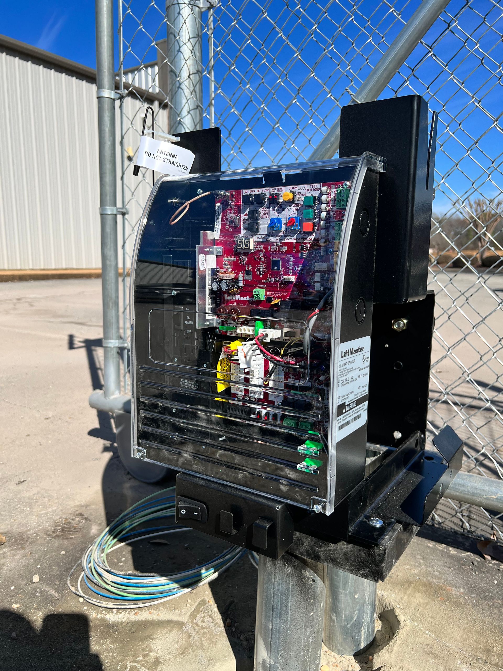 Black electrical box on a metal post next to a chain link fence; wires visible, blue sky.