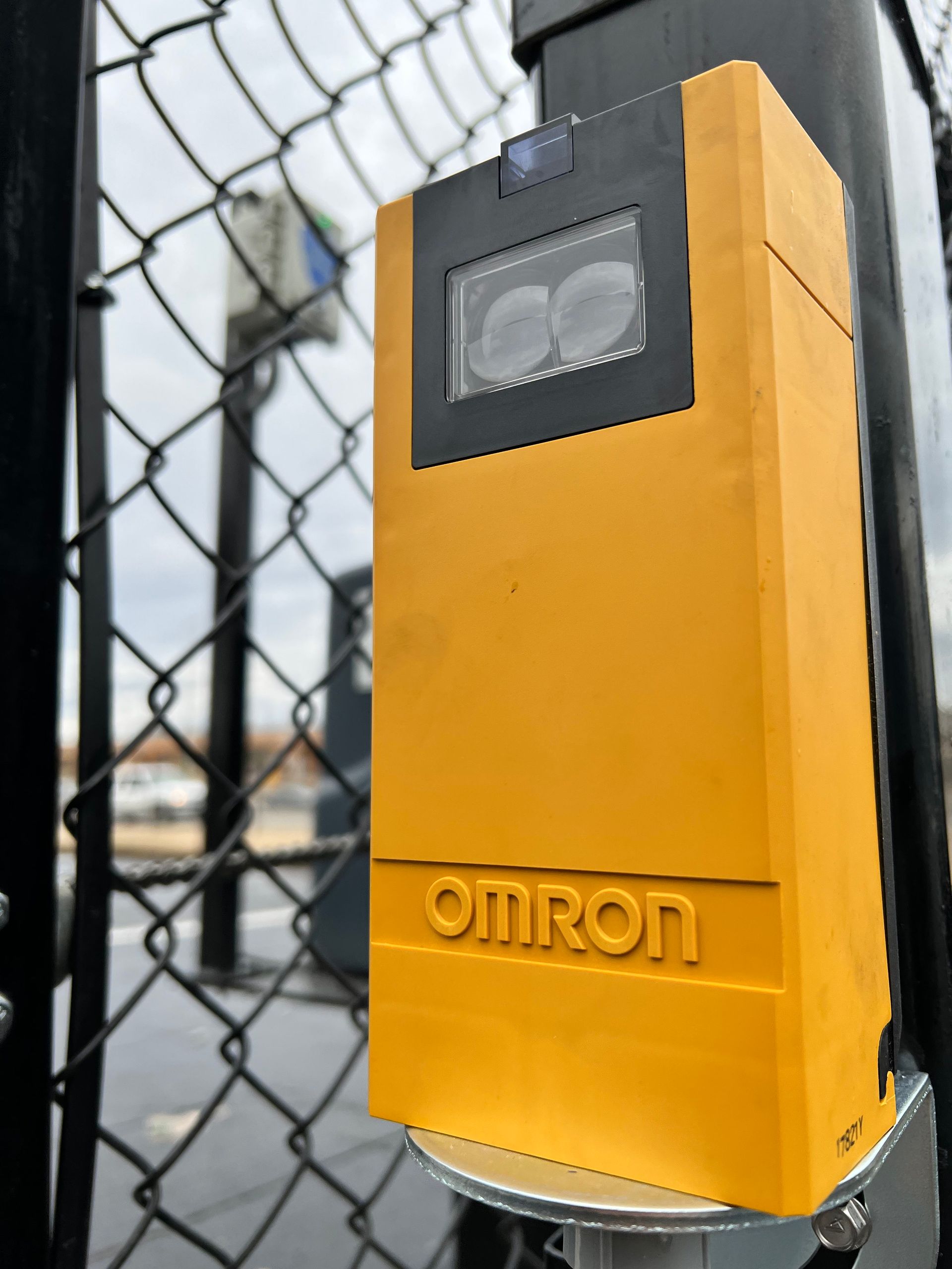 Yellow Omron sensor on a gate post; black rectangle with lenses at top. Chain link fence and sky in background.