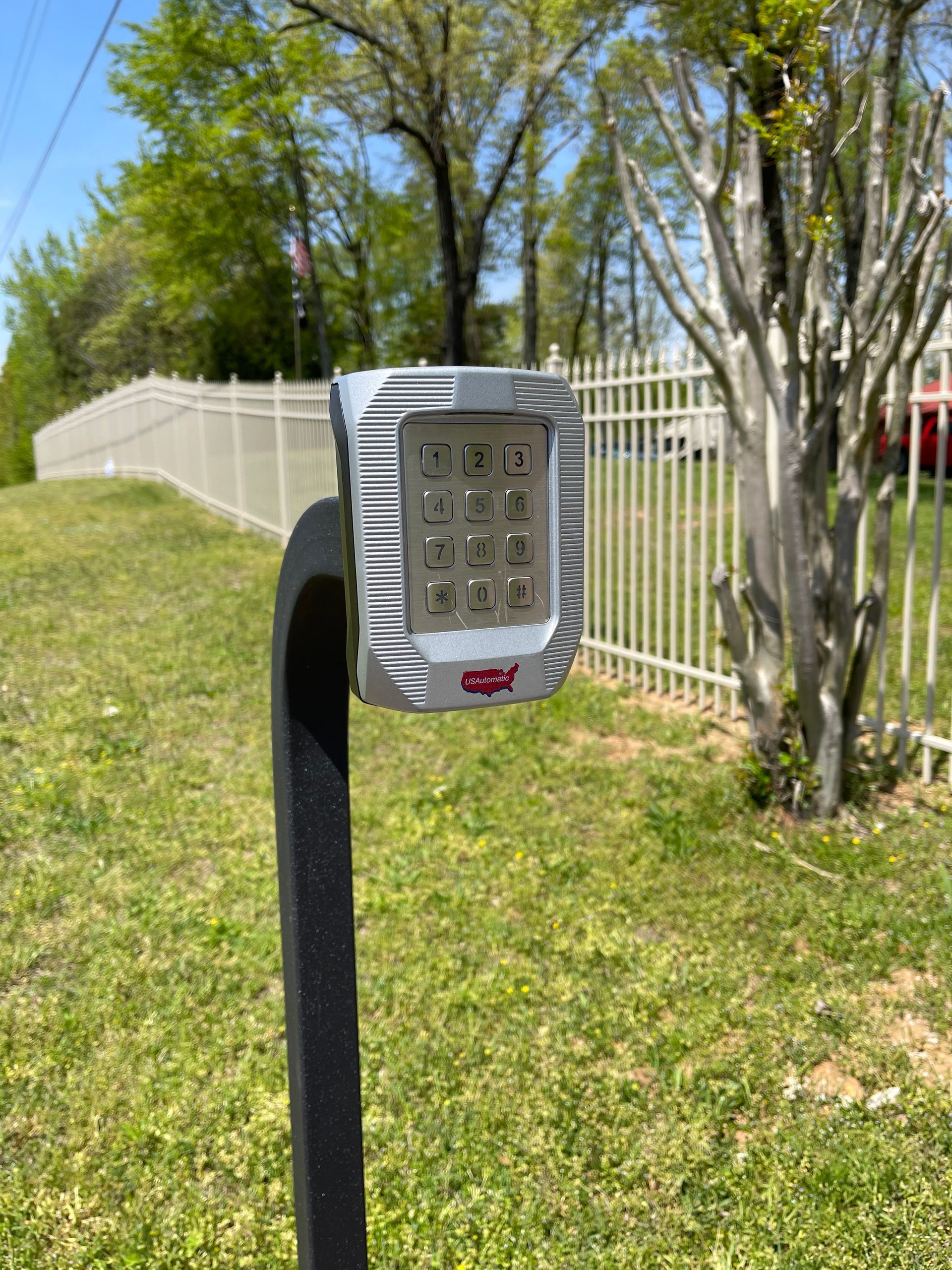 Gray LED light on a black curved post in a grassy area with a white fence and trees in the background.