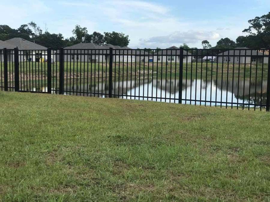 A black fence surrounds a grassy field next to a body of water.