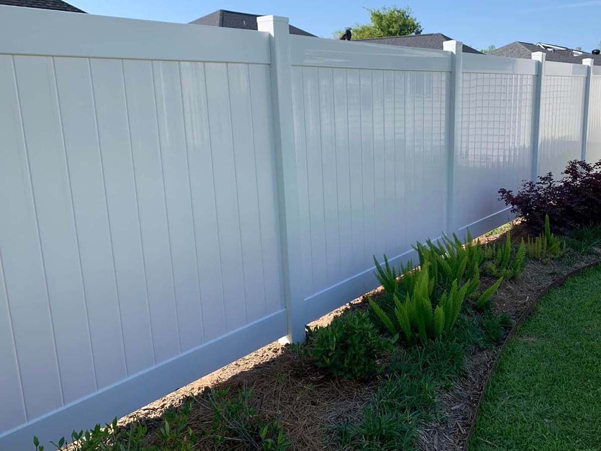 A white vinyl fence is surrounded by plants in a backyard.