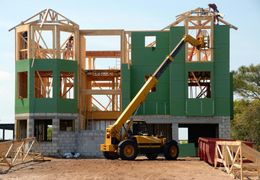 Construction site with a yellow telehandler lifting lumber for a two-story house. Green sheathing covers the wooden frame.