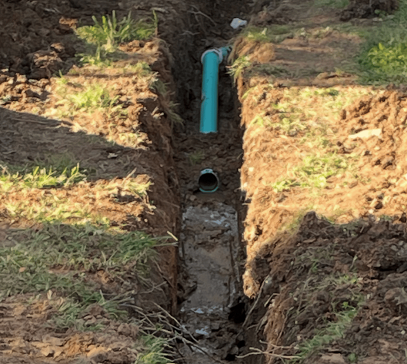 A narrow dirt trench outdoors contains a blue plastic pipe connected to a smaller grey pipe, with standing water below.