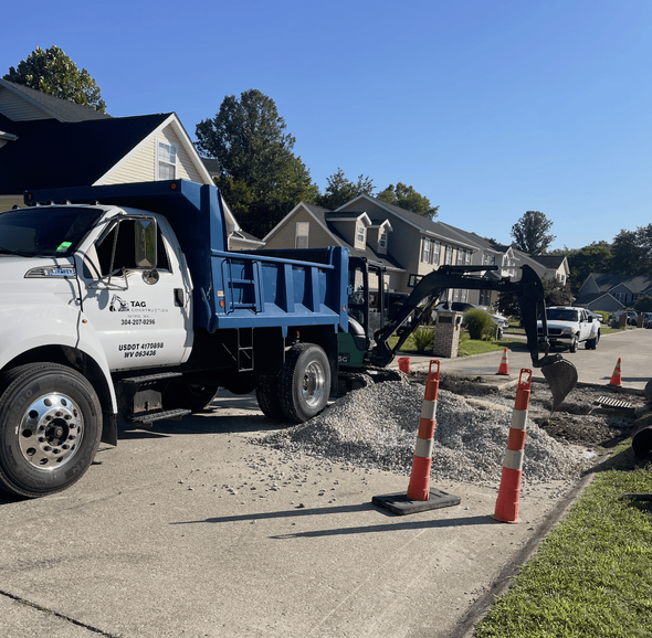 A white dump truck with a blue bed is parked on a suburban street, excavating a patch of road near orange traffic cones.